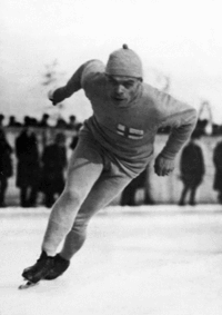 Photographie en noir et blanc d'un homme pratiquant le patinage de vitesse, le corps penché vers l'avant, jambes légèrement pliées, les bras vers l'arrière, portant sur la poitrine le drapeau finlandais.