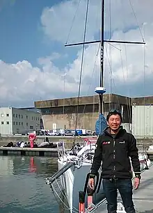 Le skipper souriant, debout devant son voilier blanc, amarré au ponton.