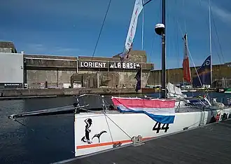 Bateau blanc amarré à un ponton, revêtu d'un drapeau néerlandais. En arrière-plan, la base de sous-marins allemande de Lorient.