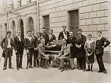 Vue en noir et blanc de 11 hommes et une femme debout derrière un piano devant lequel sont assis 2 hommes.