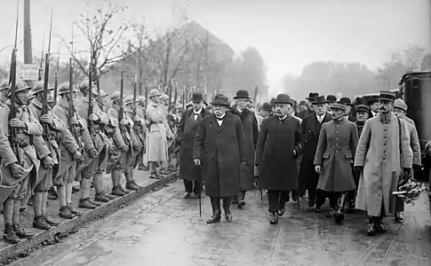 Photo en noir et blanc d’un cortège d’hommes en civil et tenue militaire passant en revue des troupes militaires