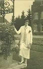 Photo en noir et blanc de Clémentine en tailleur clair portant un petit chapeau sombre, la main droit apposée sur une balustrade végétale.