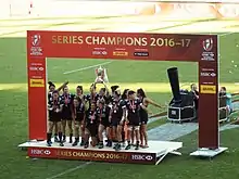 Joueuses sur un podium au centre du terrain, une joueuse tenant un trophée au dessus de sa tête.