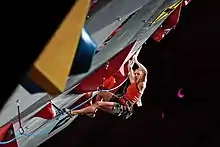 Un mur artificiel d'escalade en surplomb, à l'arrière plan la grimpeuse Petra Klingler en plein effort.