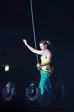 Une femme debout, avec un harnais d’escalade relié à une corde (avant ou après une ascencion), saluant de la main