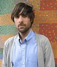  Photographie en buste d'un jeune homme aux cheveux bruns portant une chemise bleue et un gilet gris.