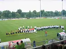 Deux équipes de football, l'une jaune et noire, l'autre vert clair, alignées sur un terrain de sport. En arrière plan : des panneaux publicitaires et des arbres