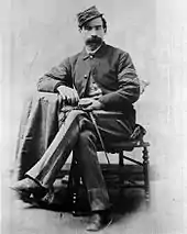 Portrait d'un homme en habits militaires, assis, portant la moustache et coiffé d'une casquette militaire.