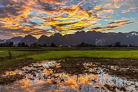 Ciel coloré avec des nuages orange et gris lumineux se reflétant dans l'eau d'une rizière, au coucher du soleil, pendant la mousson, et les montagnes de la campagne de Vang Vieng.