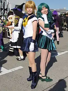 Deux jeunes filles grimées et déguisées en personnage de la série Sailor Moon. Sailor Uranus est à gauche de l’image, et a les cheveux blonds et courts, porte une robe bleue et un nœud jaune. Sailor Neptune est à droite, elle a les cheveux couleur turquoise, ils sont longs et ondulés. Son costume est bleu turquoise également.