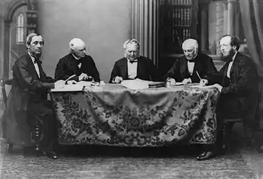 Photographie noir et blanc montrant cinq hommes en complet autour d'une table sur laquelle est déposé plusieurs liasses de document,