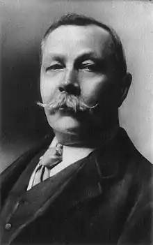 La photographie noir et blanc montre un homme d'une quarantaine d'années avec une moustache en guidon. Il porte un costume et une cravate.