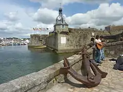 vu de l'entrée d'une ville bordée de murailles. Au premier plan est visible une ancre de grande taille.