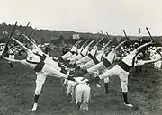 Photographie montrant des gymnastes en exercice, soulevant une barre entre les bras, une jambe relevée.