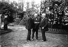 Trois hommes en costume échangeant dans un parc, avec deux individus en arrière-plan