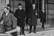 Photo en noir et blanc avec au premier plan un véhicule apparaissant flou ; deux hommes, avec chapeaux, manteaux et cravates, se trouvent sur les marches d'un bâtiment : l'homme à gauche a les cheveux blancs et une barbe grisonnante tandis que celui de droite a une moustache foncée