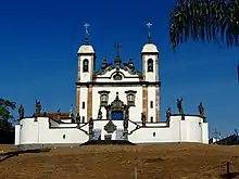 Photographie en couleurs. Vue en contre-plongée du sanctuaire avec l'esplanade et ses apôtres en premier plan, devant l'église sous un ciel bleu.