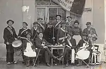 Photo noir et blanc d'une quinzaine de personnes devant une fenêtre. Les hommes ont tenues de conscrits et l'un d'entre porte un drapeau.