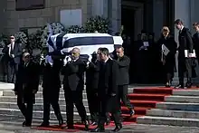 Photographie de six hommes en complet portant un cercueil recouvert d'un drapeau composé de bandes bleues et blanches.