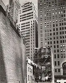 Photographie d'un petit bâtiment en briques de cinq étages où sont accrochés des cordes à linges. Le petit immeuble semble écrasé par les gratte-ciel modernes de plusieurs dizaines d'étages situés à l'arrière-plan.