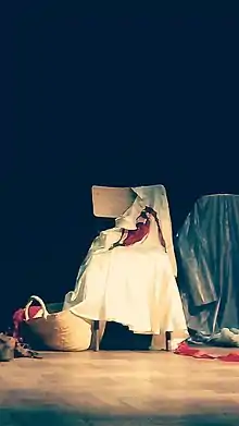 Sur une scène de théâtre, une robe blanche est posée à l'abandon sur une chaise, avec un ruban rouge. Au pied de la chaise, un panier en osier d'où sort les cheveux roux d'une poupée de chiffon.