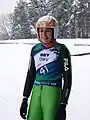 portrait jusqu'à mi-cuisses de Evelyn Insam souriante, en combinaison de saut à ski verte, coiffée de son casque, avec un dossard de compétition numéro 41, sur fond de paysage hivernal avec des sapins