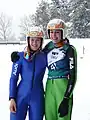 Elena Runggaldier en combinaison de saut à ski bleue, se tenant par les épaules avec Evelyn Insam en combinaison verte et dossard de compétition, toutes deux casquées et souriantes, sur fond de paysage hivernal avec des sapins enneigés