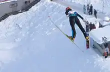 Sarah Hendrickson en combinaison noire avec un dossard, en position de vol au cours d'un saut, vu de trois quart arrière, paraissant survoler des spectateurs