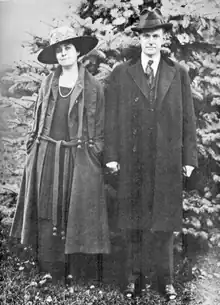 Photographie d'un homme le visage fermé tenant la main d'une femme souriante. Les deux portent une longue veste et un chapeau et la photographie a été prise en extérieur.