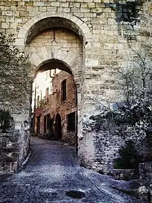 Photo d'une porte fortifiée à deux arches encadrant la rainure de la herse. A travers l'entrée, on distingue le sol, une rue pavée en montée et des maisons de pierre.