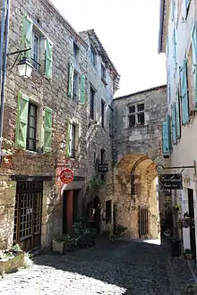 Photo couleur d'une rue pavée en pente descendante vers une porte romane. Elle est bordée de maisons en pierre.