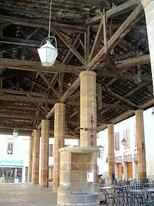 Photo couleur d'une halles à poteaux en pierre soutenant une charpente en bois. La sous-pente est grillagée contre les pigeons. La margelle d'un puits de pierre surmonté d'une croix métallique est située près d'un pilier.