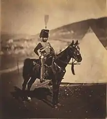 Photographie d'un homme à cheval portant un dolman et une toque en fourrure devant une tente conique.
