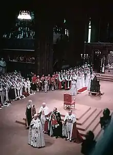 Intérieur d'une église remplie de personnes en robes et en uniformes de cérémonie. Un trône a été placé sur un piédestal au centre de l'image et la reine avec une longue traine rouge se tient à proximité.