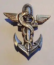 Insigne militaire porté sur la poitrine représentant une ancre de marine sur laquelle s'enroule le serpent d'Esculape, l'anneau supérieur de l'ancre est remplacé par le miroir de la sagesse