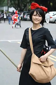 Jeune fille asiatique en robe noire unie, un nœud rouge dans les cheveux, une besace et un balai dans sa main droite.