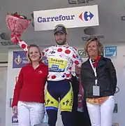 Un homme au maillot blanc à pois rouges, entouré de plusieurs personnes, soulève un bouquet de fleurs rouge.