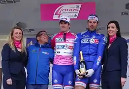 Trois hommes habillés de bleu et de rose, entourés de deux personnes, sourient, en tenant une bouteille de champagne.