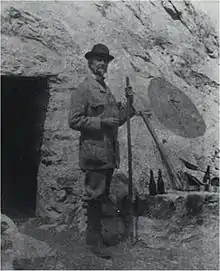 Photographie d'un homme se tenant debout devant l'entrée d'une grotte, un bâton ferré à la main.