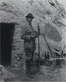 Photographie en noir et blanc d'un homme se tenant devant l'entrée d'une grotte, un bâton ferré à la main.