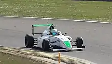 Photographie d'une monoplace de Formule 4 blanche et verte, vue de trois-quarts droit, sur une piste sèche, entourée par des bandes d'herbe.