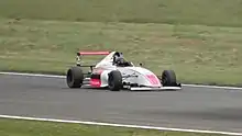 Photographie d'une monoplace de Formule 4 blanche et rouge, vue de trois-quarts droit, sur une piste sèche, entourée par des bandes d'herbe.