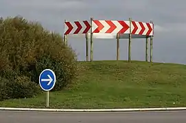 Une couronne de balises à chevrons de type K8 est ici implantée au milieu du giratoire. Non seulement il ne doit pas y avoir de balises à chevrons sur les giratoires, mais la balise K8, chevrons rouges sur fond blanc, est un équipement de signalisation temporaire.