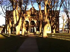 Vue partielle de la façade d'un bâtiment partiellement masqué par six arbres.