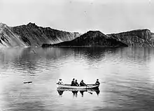 Photo en noir et blanc depuis le rivage du lac d'une barque occupée par cinq hommes devant une île volcanique conique.