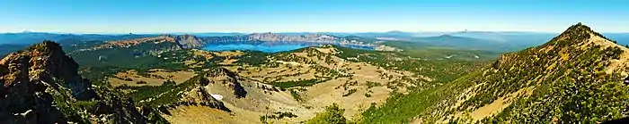 Panorama depuis un sommet vers la caldeira et son lac, dans un paysage d'éboulis, de pelouses alpines et de forêts.