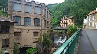  Cliché récent de l'ancienne usine du Creux de l'enfer pris depuis la rive opposée. A droite l'avenue, au centre la Durolle avec sa cascade sous un pont moderne, à gauche la façade de l'usine sur 3 niveaux. L'usine est construite pour partie sur des voûtes au dessus de l'eau, l'enduit des murs s'effritent et laisse apparaître les pierres de schistes par endroit, on distingue encore un peu le nom peint en rouge