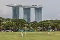 Partie de cricket avec des joueurs du cricket club au parc de Padang avec l'hôtel Marina Bay Sands en arrière plan, vu depuis les marches de la galerie nationale de Singapour. Juin 2018.