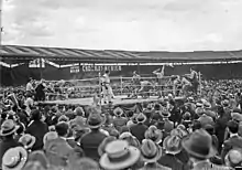 Une large foule est debout autour d'un ring de boxe sur lequel s'affrontent deux boxeurs, l'un est au sol, son coin a sauté sur le ring pour venir à son secours.
