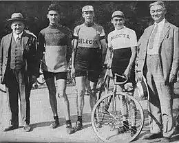 Photographie en noir et blanc montrant deux hommes en civil entourant trois cyclistes.